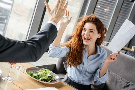 Businesspeople having business lunch at restaurant sitting eating salad drinking wine woman close-up giving five to partner holding contract happyの写真素材