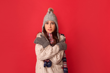 Freestyle. Young woman wearing scarf and hat standing isolated on red hugging herself feeling cold looking camera concernedの写真素材