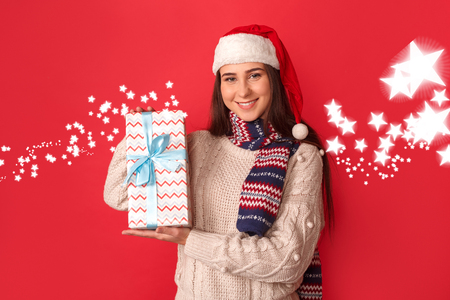 Celebration Concept. Young woman in scarf and santa hat standing isolated on red showing present to camera friendlyの写真素材