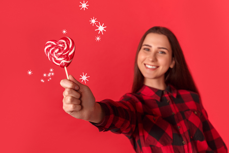 Freestyle. Young girl standing isolated on red with heart shaped lollipop close-up smiling happy blurredの写真素材