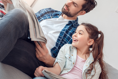Father and little daughter at home sitting girl looking at newspaper of father cheerfulの写真素材