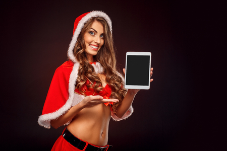 Christmas and New Year. Woman in santa costume standing isolated on black showing screen of digital tablet smiling toothyの写真素材