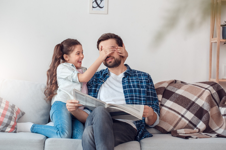 Father and little daughter at home sitting man holding newspaper girl covering his eyes playfulの写真素材