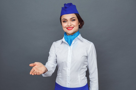 Professional Occupation. Stewardess standing isolated on grey giving hand towards camera smiling friendlyの写真素材