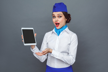 Professional Occupation. Stewardess standing isolated on grey showing screen of digital tablet looking camera surprisedの写真素材