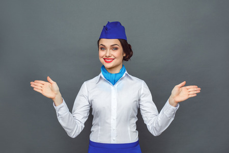Safety Regulations. Stewardess standing isolated on grey showing emergency exit aside looking camera joyfulの写真素材