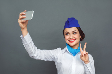 Professional Occupation. Stewardess standing isolated on grey taking selfie on smartphone showing peace sign smiling playfulの写真素材