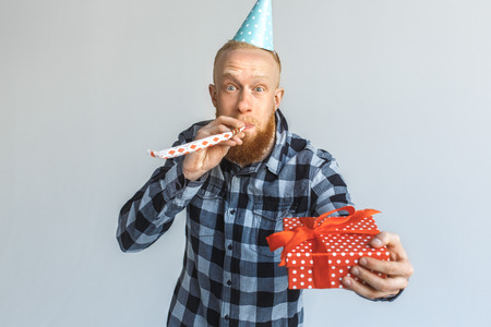 Birthday Concept. Mature man in cap standing isolated on grey giving gift to camera blowing party horn cheerfulの写真素材