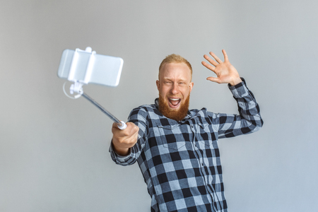 Mature man standing isolated on grey with mono pod taking selfie on smartphone waving to camera friendlyの写真素材