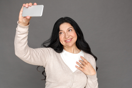 Mature lady standing isolated on grey taking selfie on smartphone smiling cheerfulの写真素材