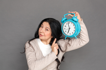 Freestyle. Mature lady standing isolated on grey shwoing time on alarm clock concernedの写真素材