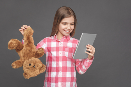 Freestyle. Girl standing isolated on grey with teddy bear indifferent looking at digital tablet excitedの写真素材