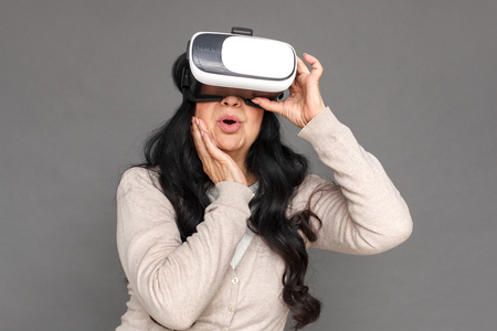 Freestyle. Mature lady in virtual reality headset standing isolated on grey watching video impressedの写真素材