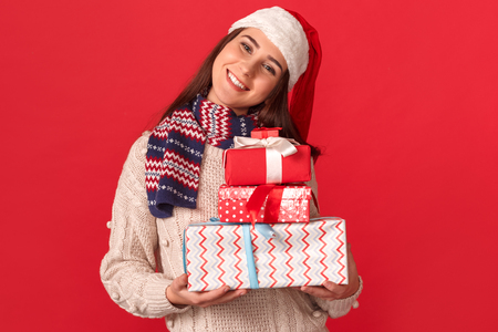 Celebration Concept. Young woman in scarf and santa hat standing isolated on red with gift boxes smiling joyful close-upの写真素材