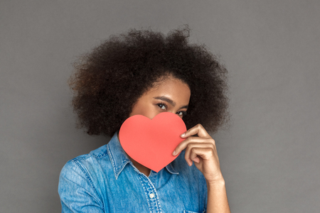 Freestyle. Mulatto woman standing isolated on grey covering face with heart shape card smiling playful close-upの写真素材