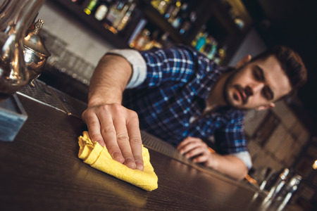 Young bartender standing at bar counter cleaning surface blurred backgroundの写真素材
