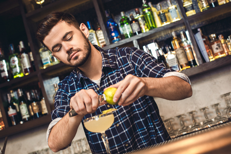Young bartender standing at bar counter cutting lime zest into coktail concentratedの写真素材