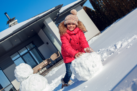 Winter vacation. Girl standing outdoors near house rolling snowball making snowman smiling happyの写真素材