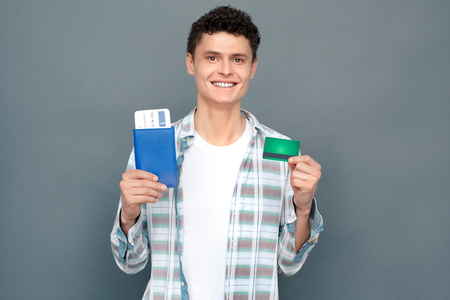 Man isolated on gray wall tourism concept standing holding passport with ticket and credit card smiling cheerfulの写真素材