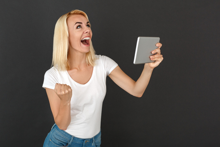 Freestyle. Woman standing isolated on black wall with digital tablet looking up laughing happy successfulの写真素材
