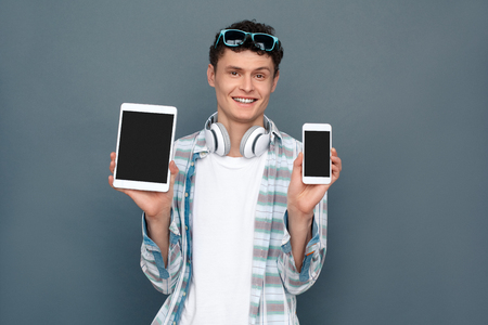 Man isolated on gray wall tourism concept standing holding smartphone and digital tablet looking camera cheerfulの写真素材