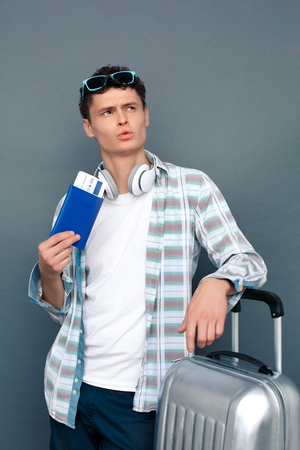 Man isolated on gray wall tourism concept standing leaning on suitcase holding passport with ticket looking aside thoughtfulの写真素材