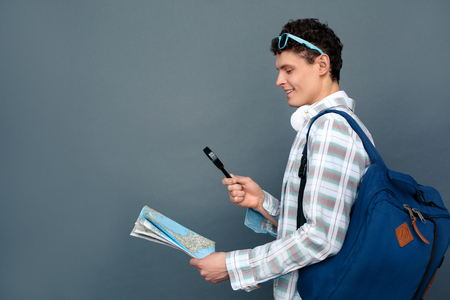 Man isolated on gray wall tourism concept walking with backpack magnifying map happyの写真素材