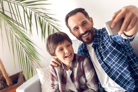 Father and little son at home sitting on sofa taking selfie on phone looking camera laughing joyfulの写真素材