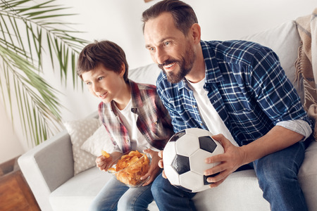 Father and little son at home sitting on sofa boy eating potato chip watching soccer match together with dad holding ball looking at screen cheerfulの写真素材