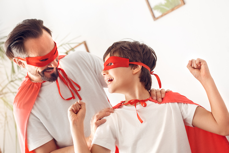 Father and son in superheroes costumes at home standing looking at each other laughing cheerfulの写真素材