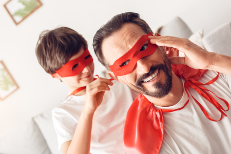 Father and son in superheroes costumes at home boy helping tying mask to dad smiling close-upの写真素材
