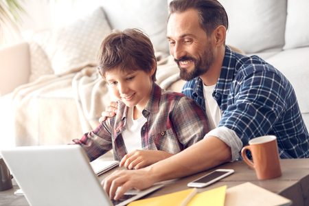 Father and son at home sitting at table playing game on laptop smiling happyの写真素材