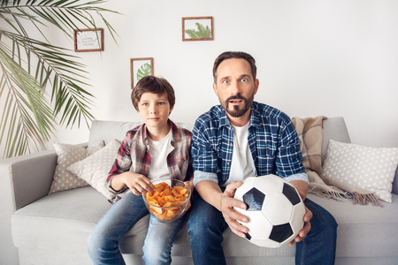 Father and little son at home sitting on sofa boy eating potato chip dad with ball watching soccer looking camera impressedの写真素材