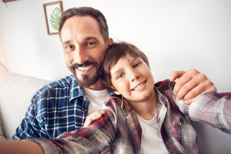 Father and little son at home sitting hugging taking selfie on smartphone looking camera happy close-upの写真素材