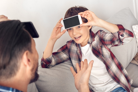 Father and little son at home sitting on sofa playing heads up with smartphone boy close-up looking at dad excitedの写真素材