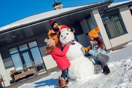 Winter vacation. Family time together outdoors standing near house making snowman smiling concentratedの写真素材