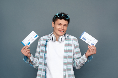 Man isolated on gray wall tourism concept standing holding two tickets choosing destination confusedの写真素材