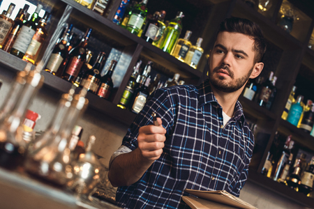 Young bartender standing at bar counter holding pen and notebook thoughtfulの写真素材