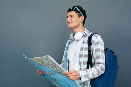 Man isolated on gray wall tourism concept walking with backpack holding map looking forward cheerfulの写真素材