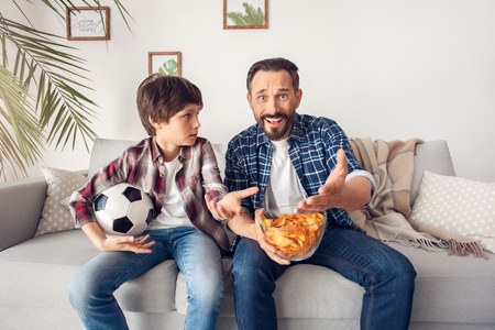 Father and little son at home sitting on sofa holding bowl of chip and ball watching soccer pointing at screen swearingの写真素材