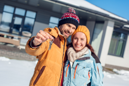 Winter vacation. Young couple standing together outdoors with keys from new house close-up smiling cheerful blurredの写真素材