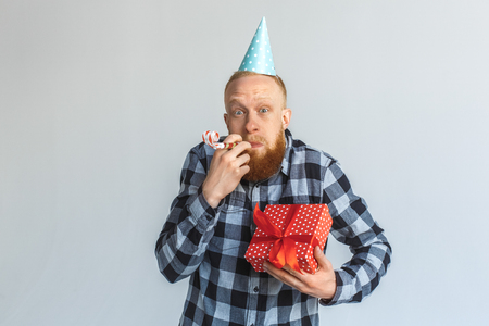 Birthday Concept. Mature man in cap standing isolated on grey with gift party horn in mouth looking camera playfulの写真素材
