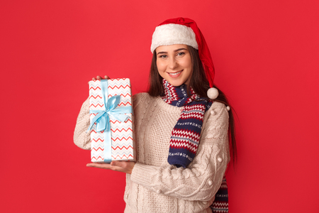 Celebration Concept. Young woman in scarf and santa hat standing isolated on red showing present to camera friendlyの写真素材