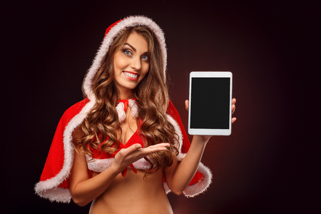 Christmas and New Year. Woman in santa costume standing isolated on black showing screen of digital tablet smiling playfulの写真素材