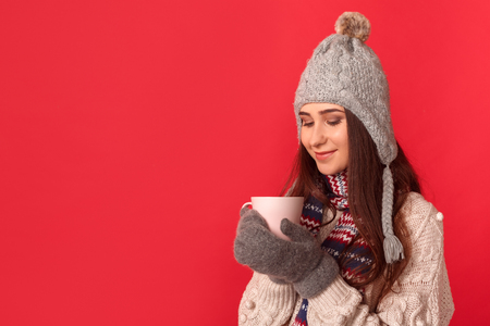 Freestyle. Young woman wearing scarf and hat standing isolated on red looking at cup of cocoa smiling joyfulの写真素材
