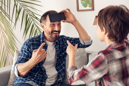 Father and little son at home sitting on sofa playing heads up with smartphone guessing word cheerfulの写真素材