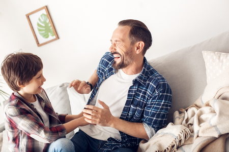 Father and little son at home sitting on sofa boy tickling dad laughing cheerfulの写真素材
