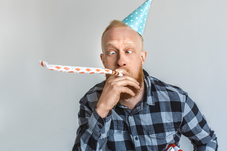 Birthday Concept. Mature man in cap standing isolated on grey blowing into party hor looking at nose silly close-upの写真素材