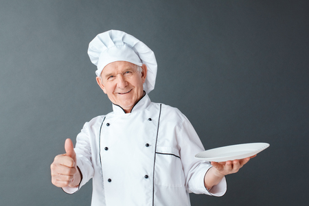 Senior chef studio standing isolated on gray with plate looking camera playful thumb upの写真素材