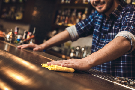 Young bartender standing at bar counter wiping table with rag happy close-upの写真素材